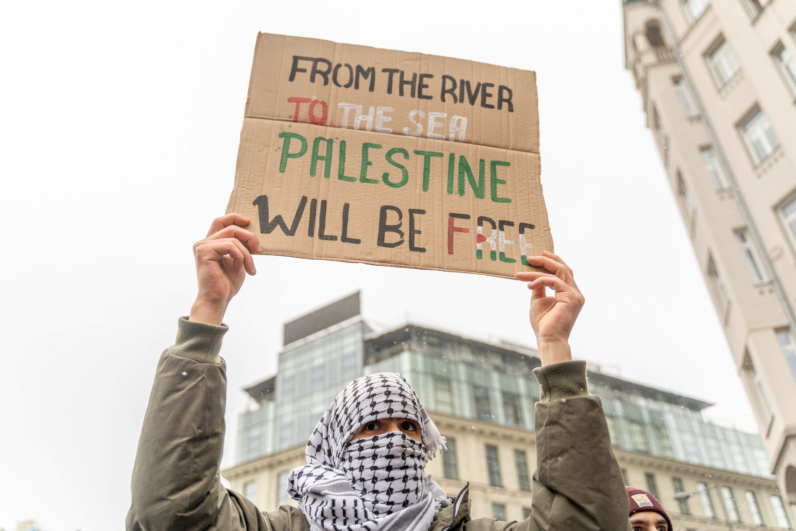 a person with a keffiyeh covering their face, with only their eyes visible, holds a homemade cardboard sign above their head on which is written in the black, white, red and green colours of the palestinian flag: 'from the river to the sea, palestine will be free'